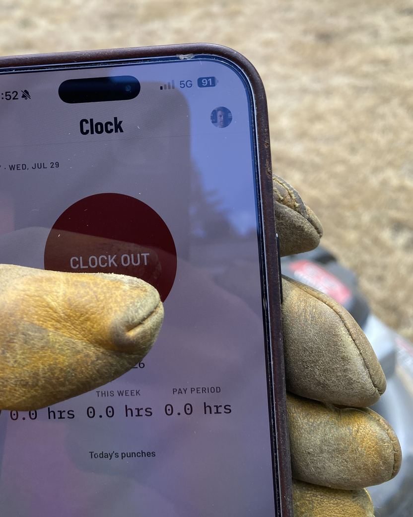A work-gloved hand holding a phone running CutCrew&rsquo;s Clock screen mid-job: a red CLOCK OUT button above the day&rsquo;s hours.